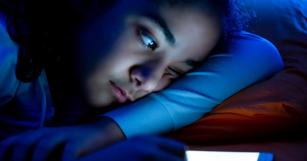 A young woman lying on her side, with the reflection of the blue light of her mobile device shining on her
