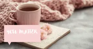A pink mug of tea nestles near a shawl on a table, adorned. Beside it, a thought bubble : you matter.